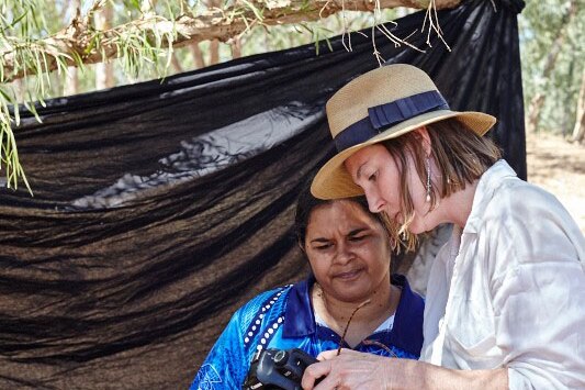 Two women examine the settings on a camera in front of a makeshift black backdrop in the bush.