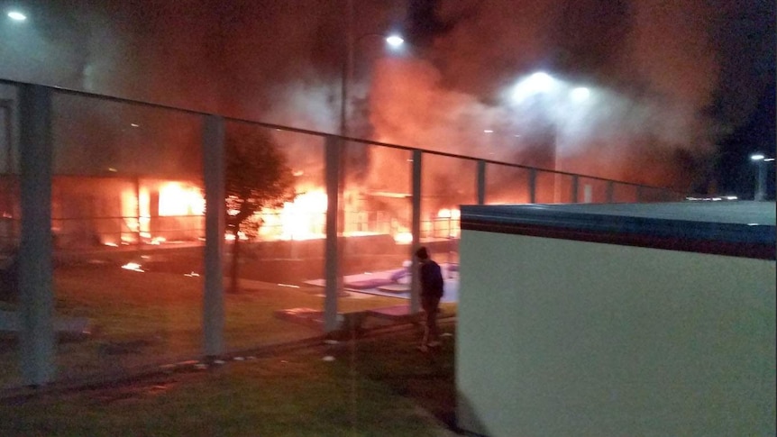 Buildings burn and a man watches through a metal fence.