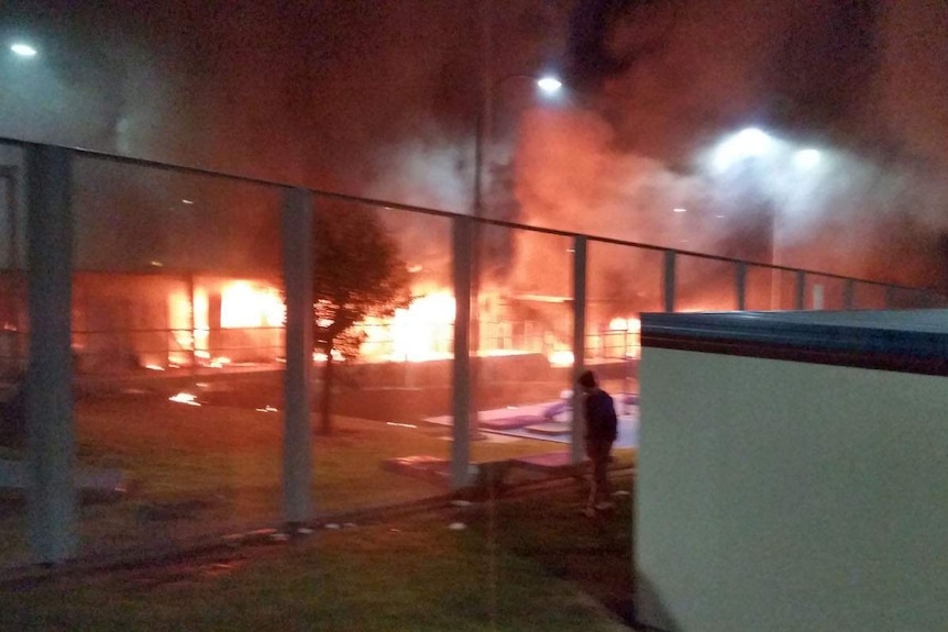 Buildings burn and a man watches through a metal fence.