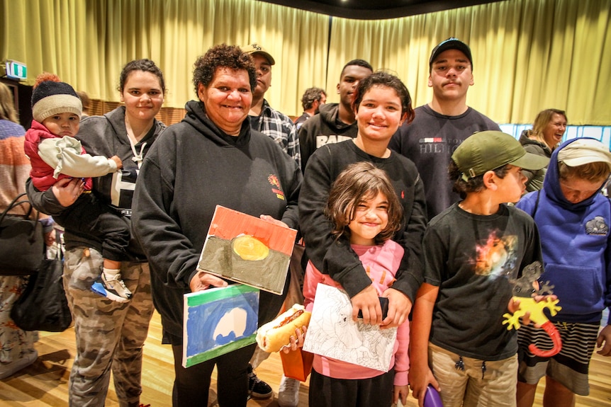 A woman with her kids holds an Aboriginal flag drawing in one hand and a Torres Strait Islander drawing in the other