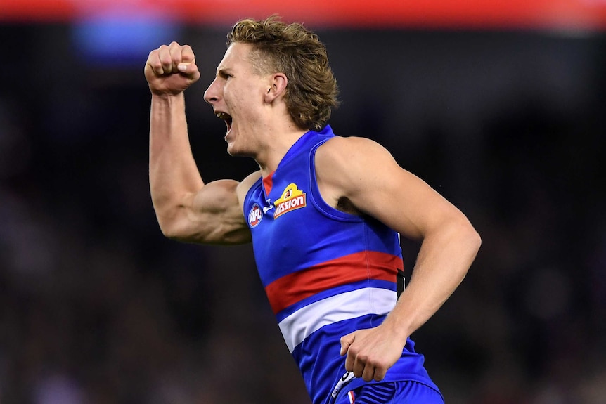 A male AFL player raises his right fist as he screams out in celebration after kicking a goal.