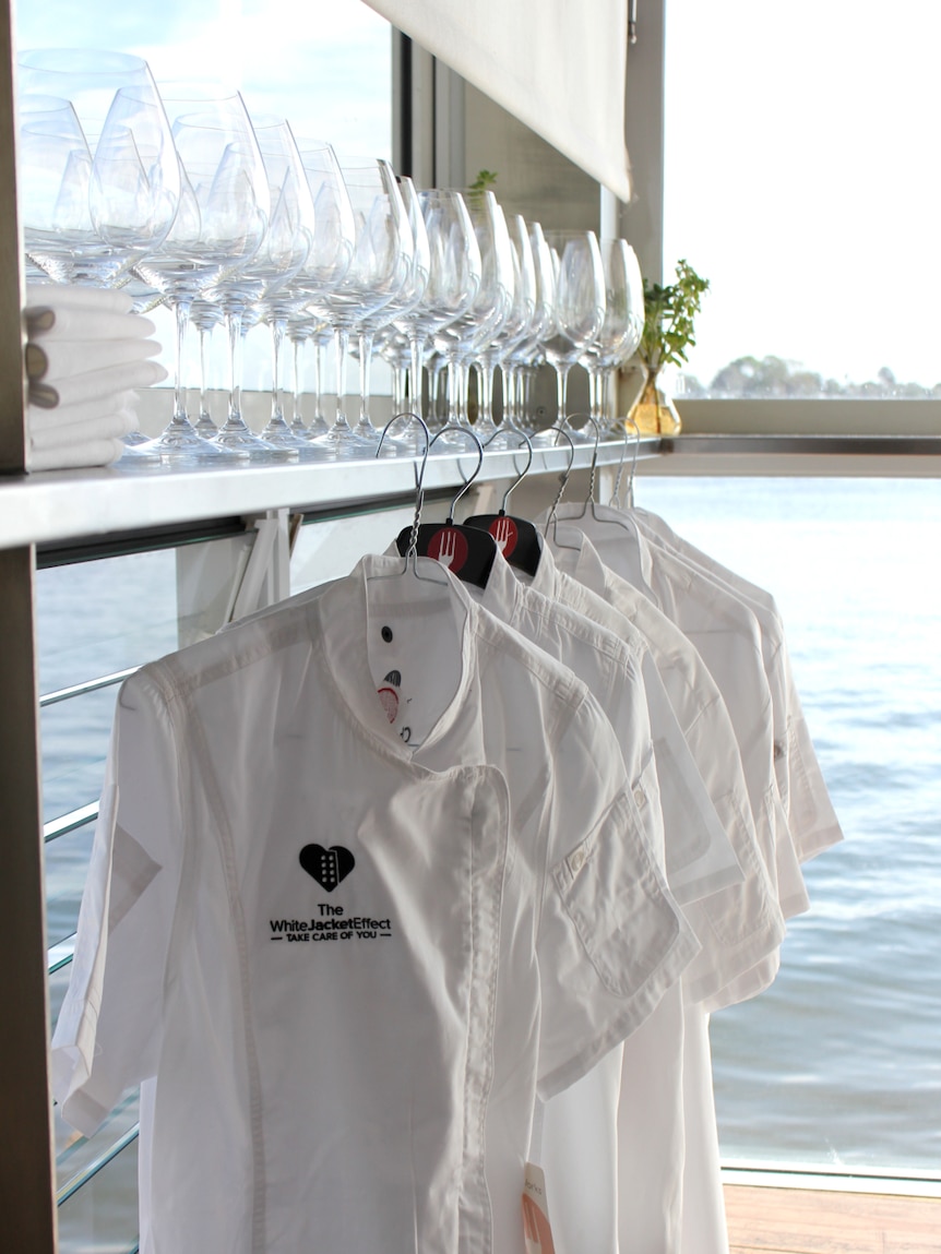 White chef's jackets hang on a bar with wine glasses and napkins in a waterfront restaurant.