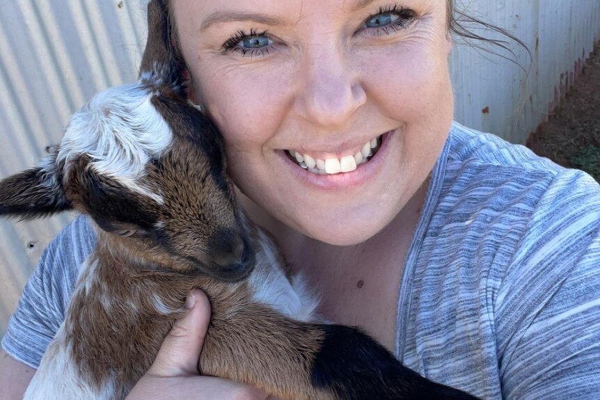 Woman grins as she cradles a small goat with their faces side by side