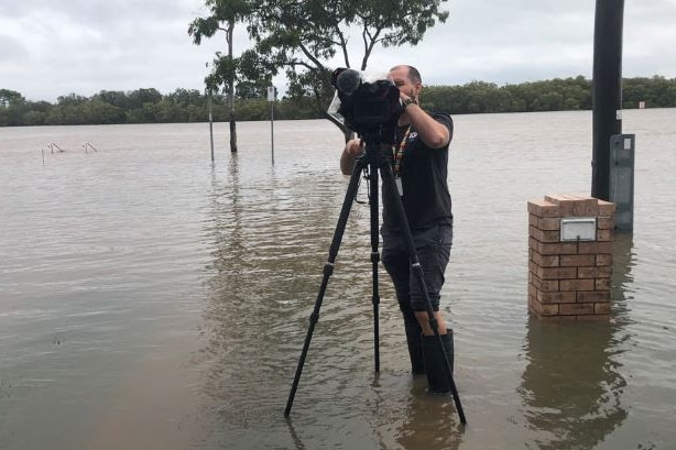Cameraman looking through camera on tripod while standing in ankle deep water, wearing gumboots.