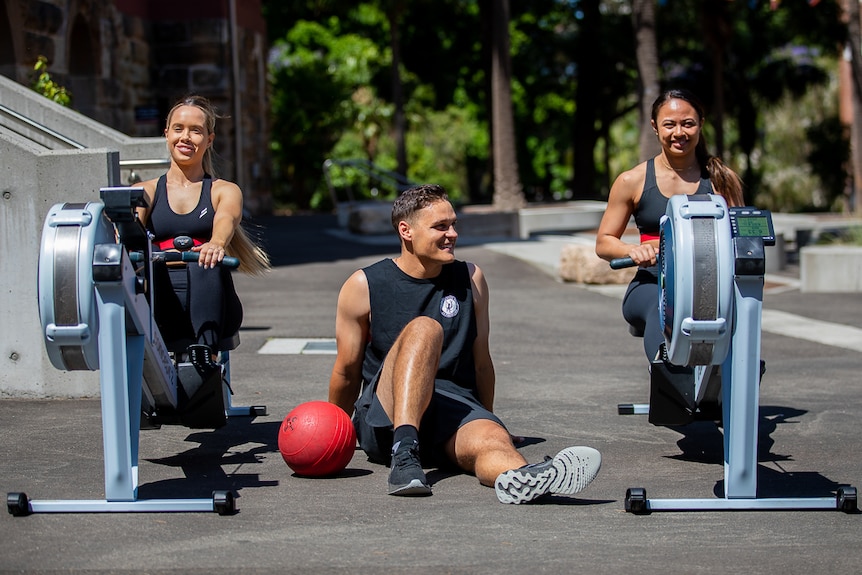 Two women on rowing machines. One man sitting in between the machines with a fitball next to him.