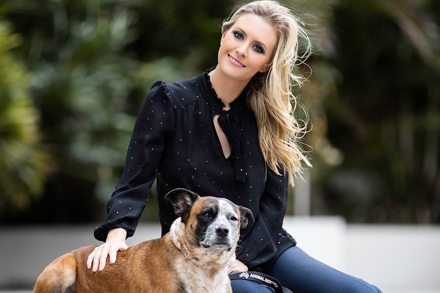 A woman with long blonde hair sitting on the ground with a medium sized mostly brown dog.