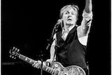 Black and white photo of Paul McCartney playing live and hold one arm aloft 