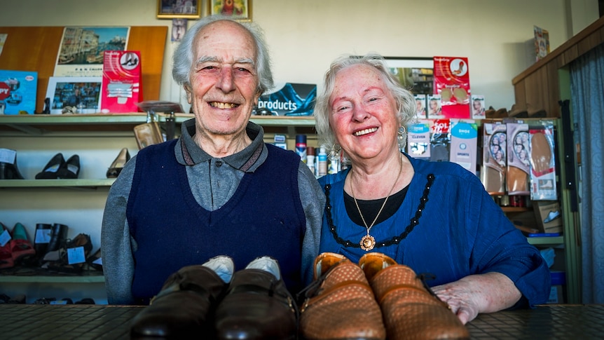 An elderly couple smile at the camera with two pairs of shoes sitting on a bench in front of them