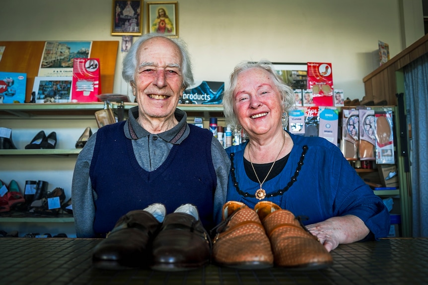 An elderly couple smile at the camera with two pairs of shoes sitting on a bench in front of them