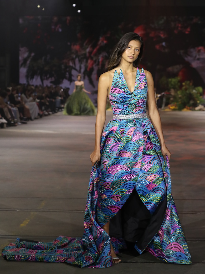 Indigenous model wears vibrant bright couture dress that is blue, pink, purple and yellow on the fashion runway.