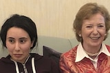 A young woman with dark hair, wearing a dark tracksuit jumper, sits on a green couch next to an older woman smiling