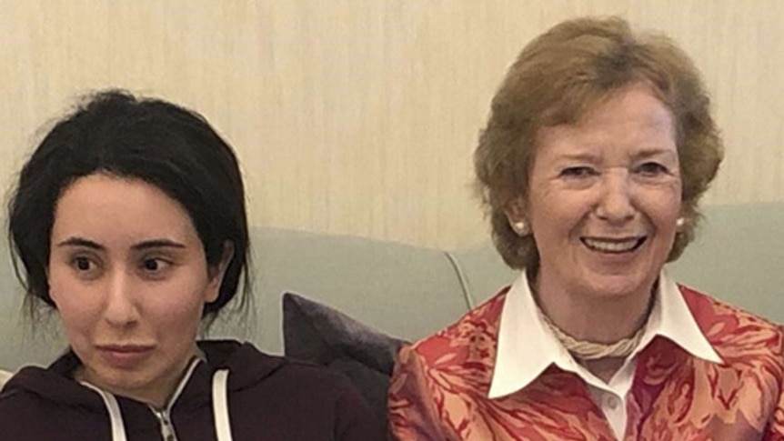 A young woman with dark hair, wearing a dark tracksuit jumper, sits on a green couch next to an older woman smiling