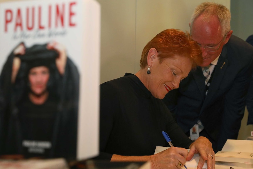 Pauline Hanson signs her autobiography and smiles as Steve Dickson speaks into her left ear