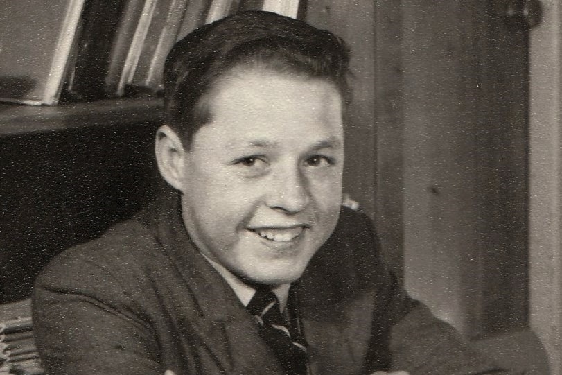 A schoolboy in uniform with blazer and tie smiles at the camera in a black and white photo