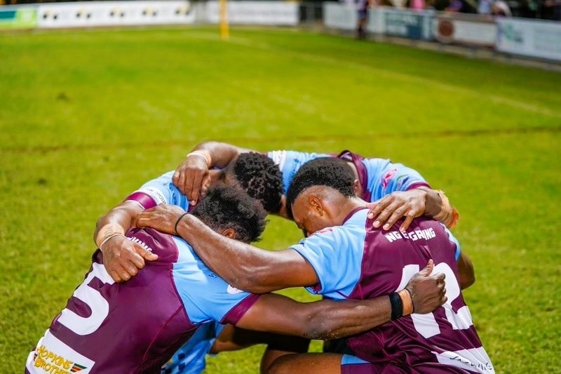 Four men from Papua New Guinea wearing maroon and sky blue football jerseys kneeling on a field and praying.