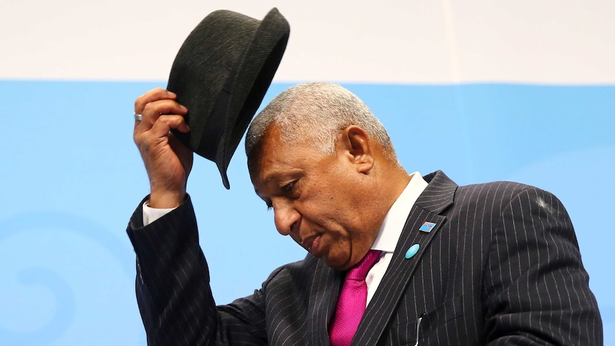 Frank Bainimarama takes off his hat. He is wearing a dapper pinstripe suit with bright magenta tie.