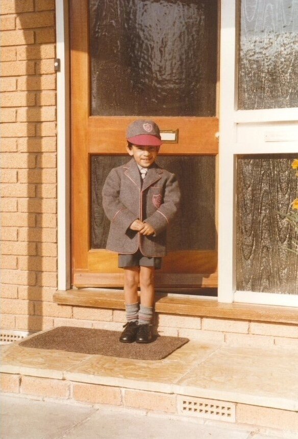 Ένα μικρό αγόρι με γκρι και κόκκινη σχολική στολή σφίγγει τα χέρια του ενώ ποζάρει για μια εικόνα σε μια εξώπορτα ενός σπιτιού