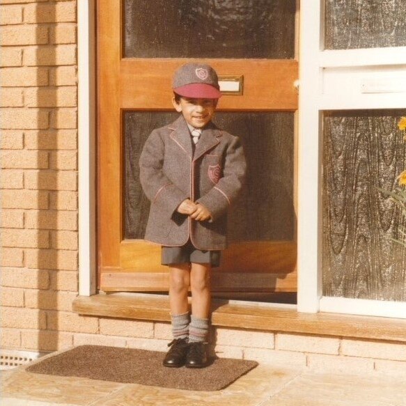 Ένα μικρό αγόρι με γκρι και κόκκινη σχολική στολή σφίγγει τα χέρια του ενώ ποζάρει για μια εικόνα σε μια εξώπορτα ενός σπιτιού
