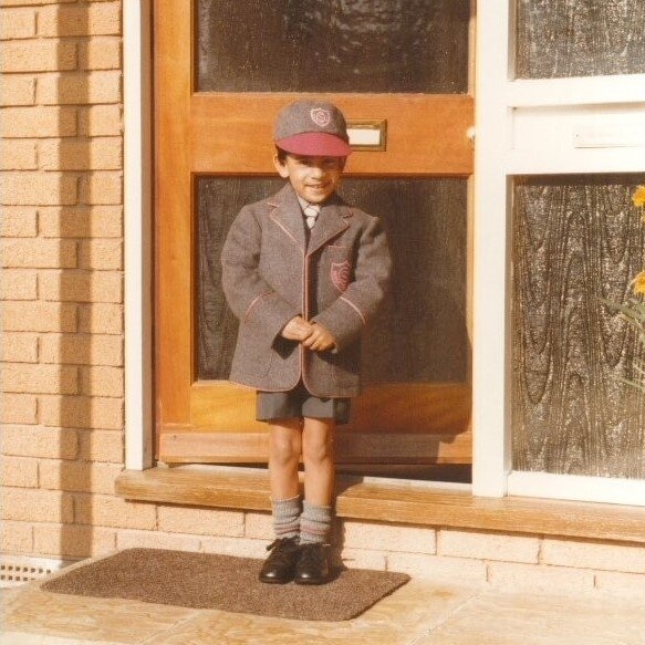 A little boy in a grey and red school uniform clasps his hands while posing for an image at a front door of a house