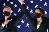 Joe Biden and Kamala Harris raise their hands together in front of a US flag while a firework exploded behind them