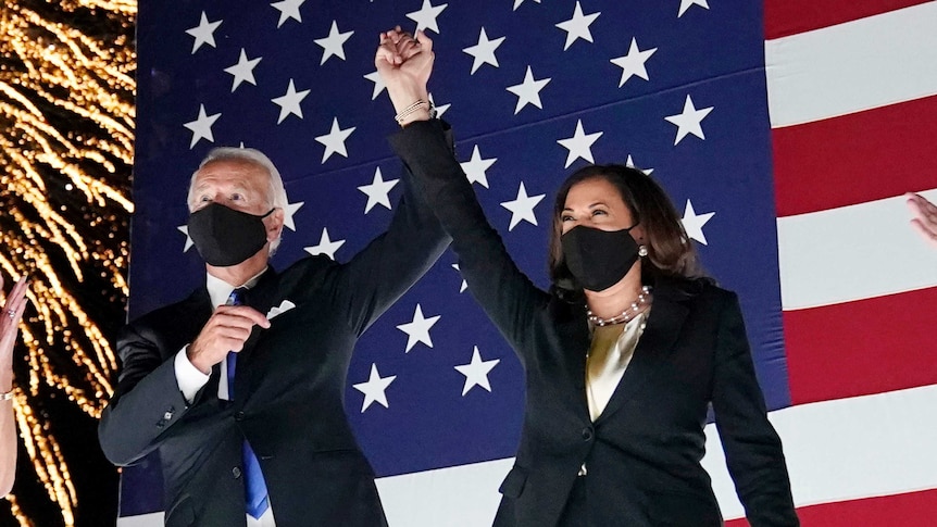Joe Biden and Kamala Harris raise their hands together in front of a US flag while a firework exploded behind them