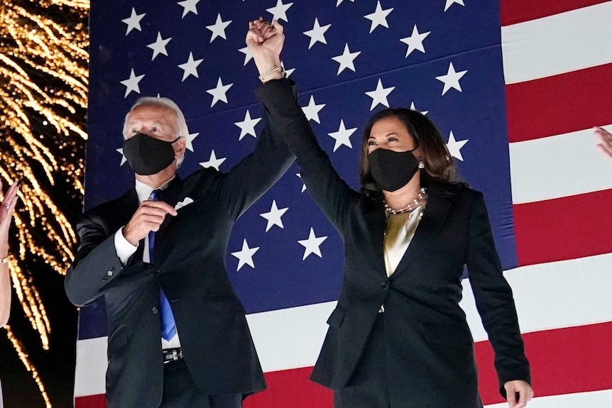 Joe Biden and Kamala Harris raise their hands together in front of a US flag while a firework exploded behind them