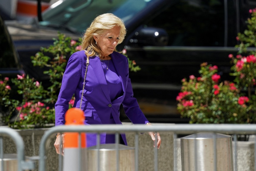Jill Biden, wearing a purple suit, walks behind a barricade. There are flowers in planters behind her.