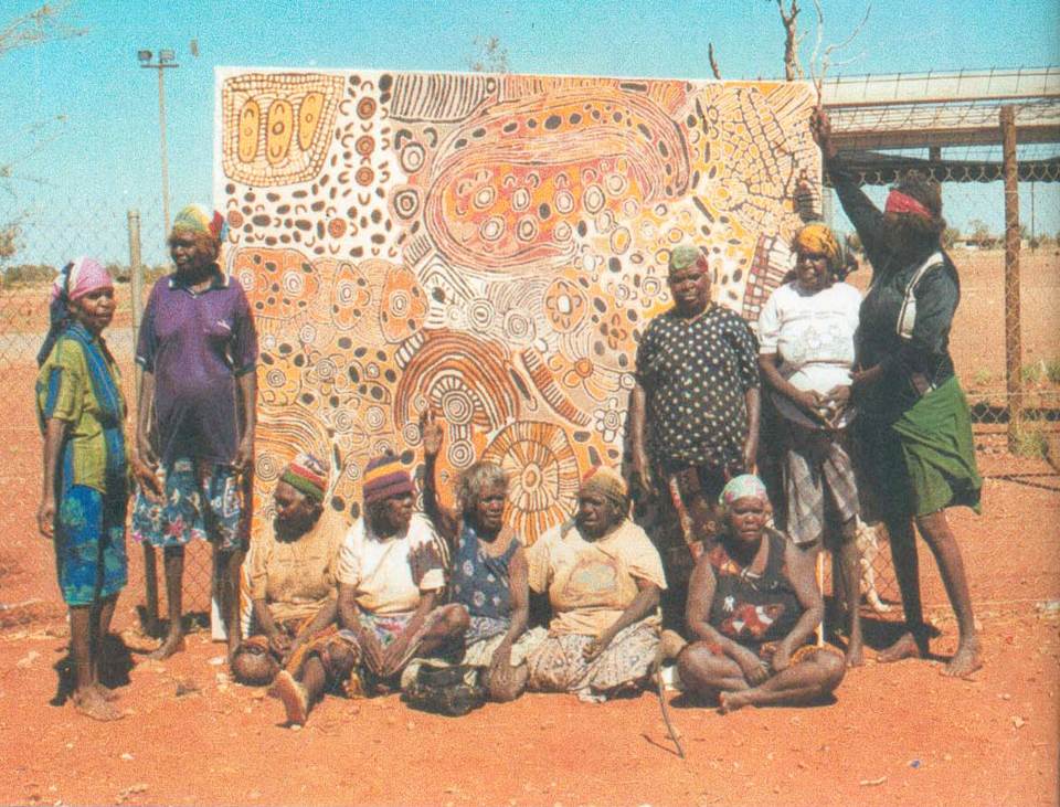 A group of Aboriginal women stand and sit around a large painting in the desert.