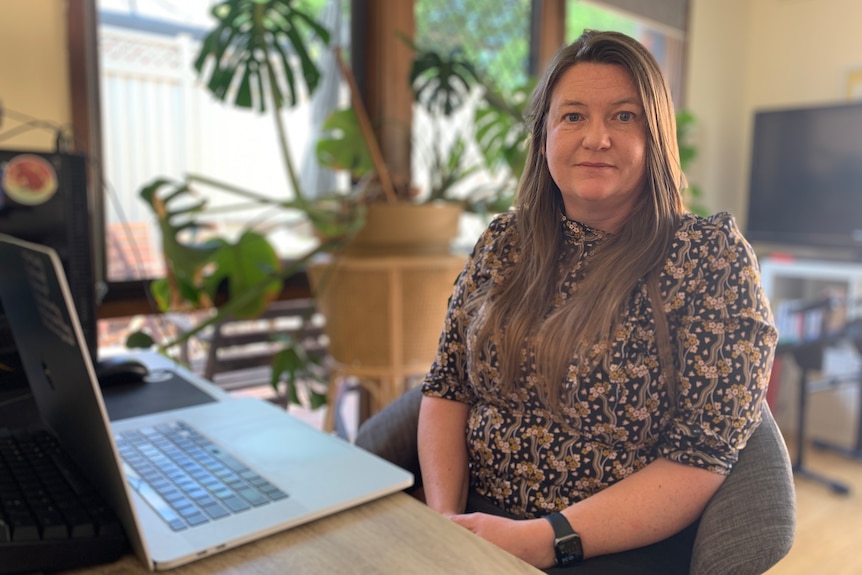 A woman sitting at a laptop. She has long brown hair and is wearing a floral blouse