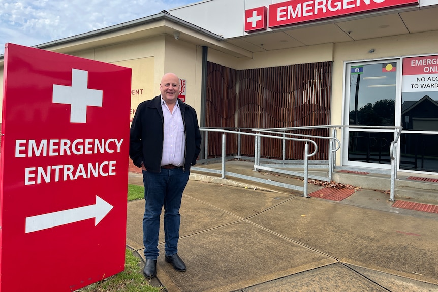 A man stands outside a hospital emergency department, next to a large red sign
