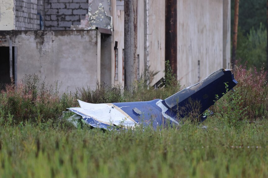 A destroyed fragment of a plane's body sitting in a field in front of a dilapidated structure