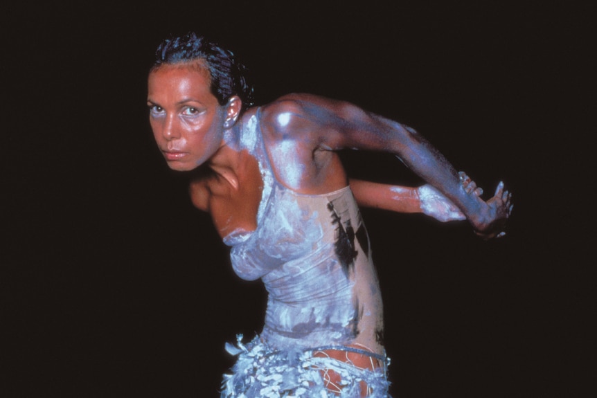 An Indigenous woman in her 40s in a white body paint and feathers, dancing