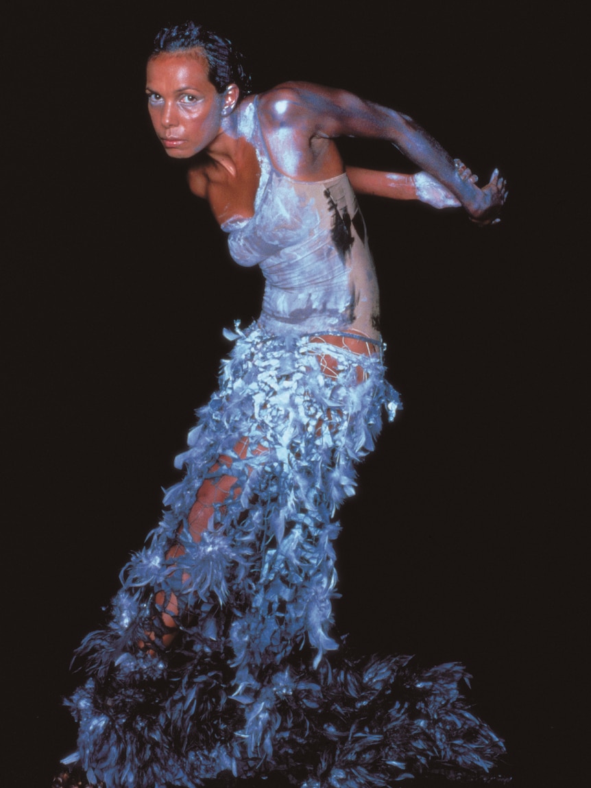 An Indigenous woman in her 40s in a white body paint and feathers, dancing