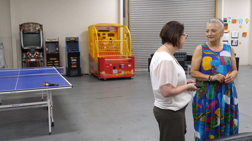 two women standing in a warehouse, with arcade games and a table tennis table