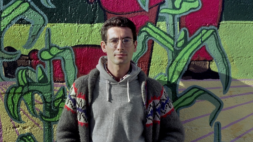A white man with short brown hair and glasses, wearing a grey hoodie and a cardigan, stands against a graffitied wall