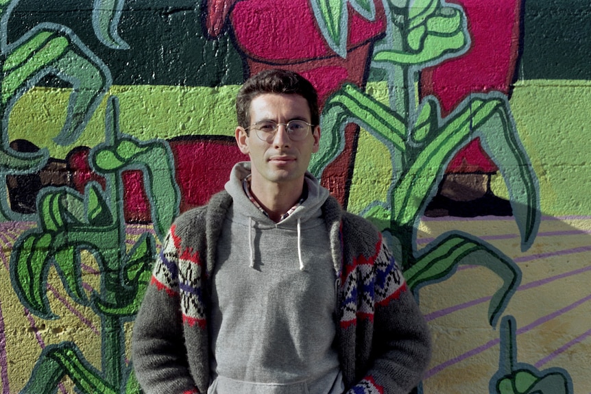 A white man with short brown hair and glasses, wearing a grey hoodie and a cardigan, stands against a graffitied wall