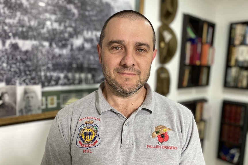 A man looks at the camera. He is wearing a grey polo shirt with a poppy embroidered. There is war memorabilia in the background