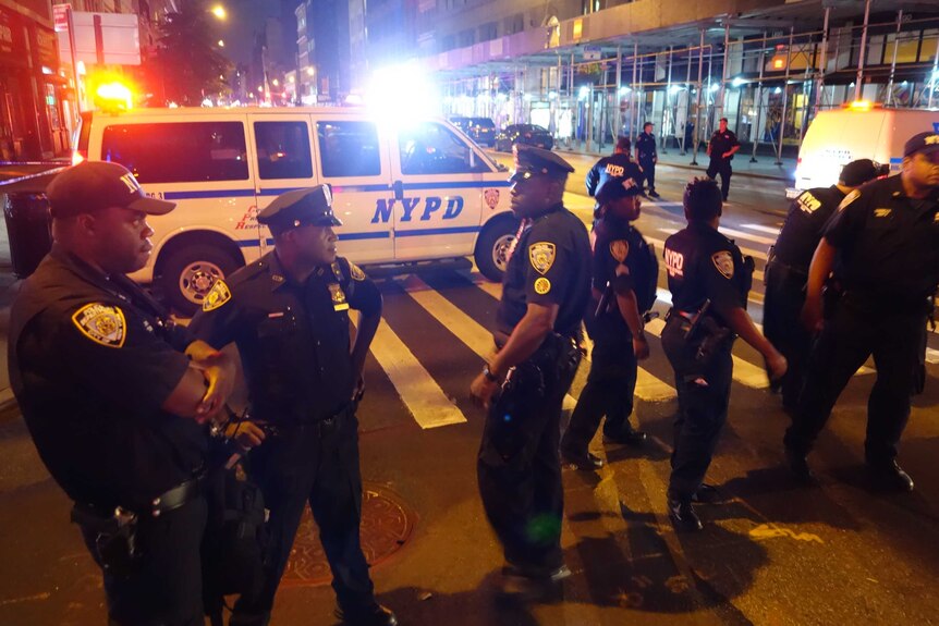 A large group of New York police stand on a street. In the background is a police vehicle with its emergency lights on.