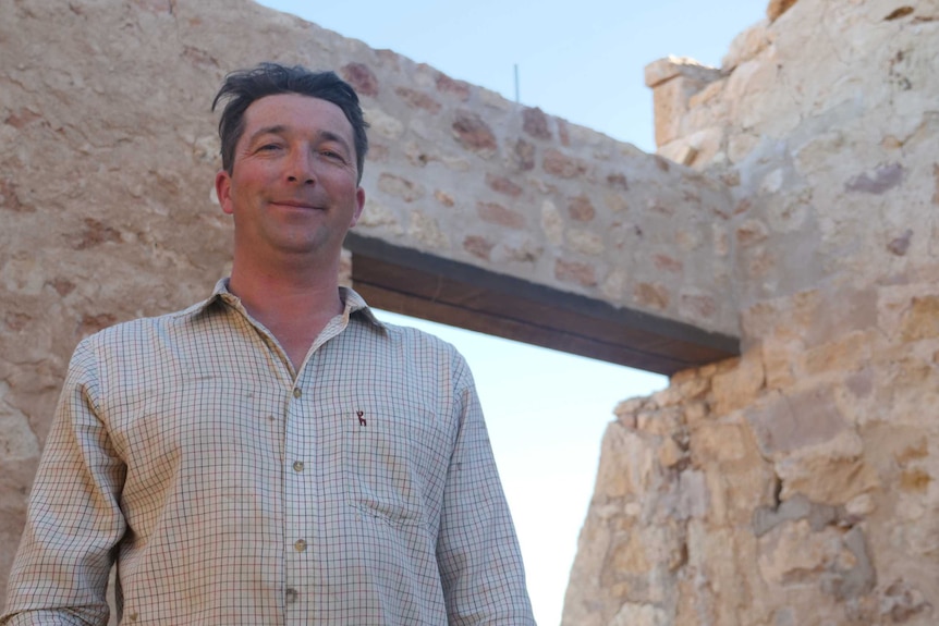 A man with dark brown hair in a white stripy shirt stands next to a wall built of stone.