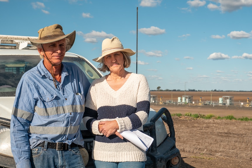 A farming couple stand near a farm ute with gas wells behind them,