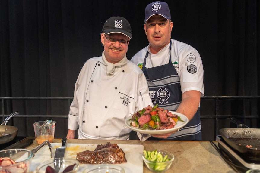 Two chefs hold a plate of meal featuring meat.