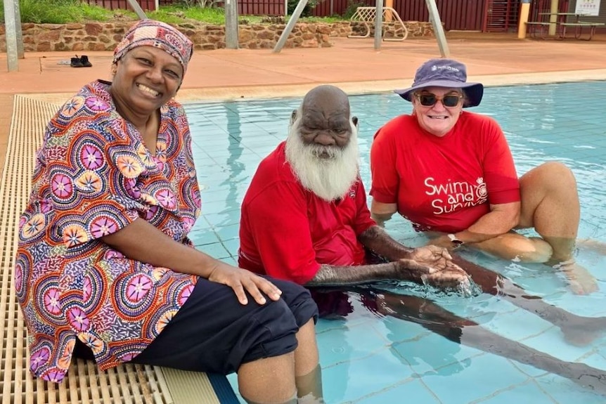 a woman and a man sit in the shallows while another woman smiles sitting on the pool edge