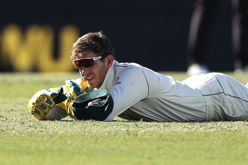 Australia wicketkeeper Tim Paine lies on the grass during a Test cricket match. His sunglasses are slightly askew.