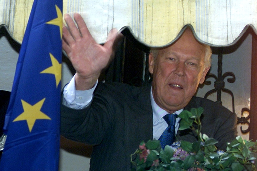 A man dressed in a dark suit looks out from a window waving next to a European Union flag.