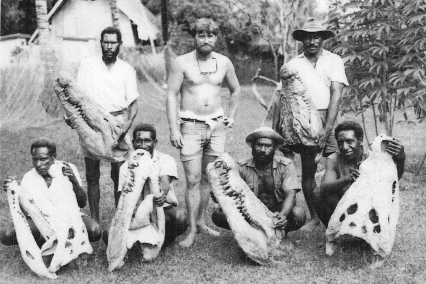 A shirtless Caucasian man standing in the middle of six Papua New Guinean men each holding large crocodile skulls.