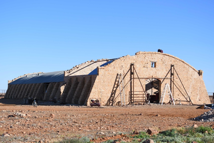 A huge, long stone building with half of its roof missing stretches into the distance on red dirt.