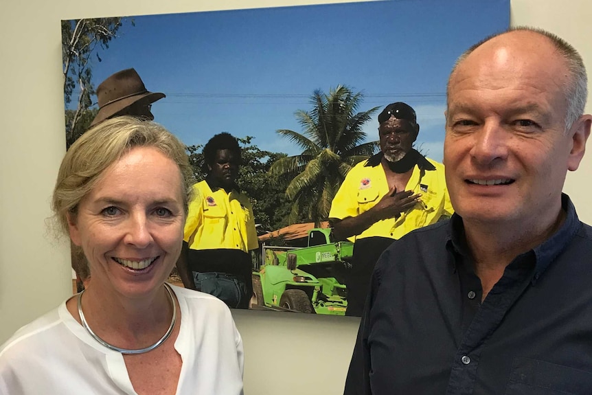 A woman in a white shirt and a man in a black shirt standing in front of a photo of two employees.