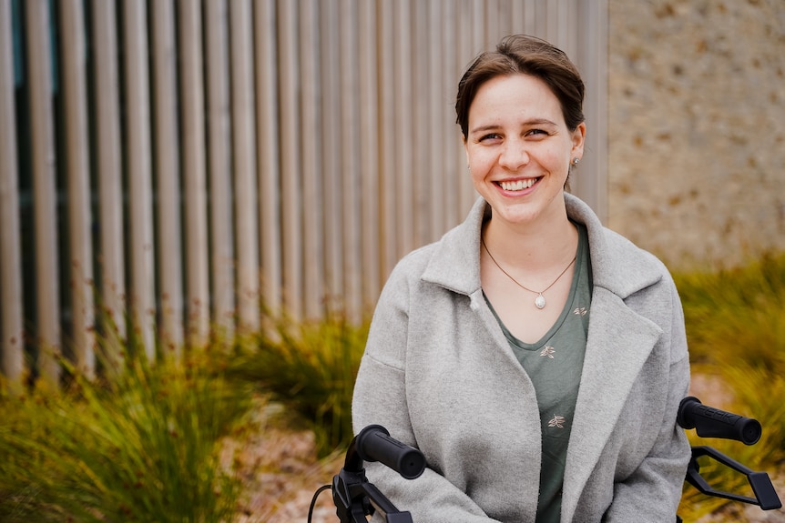 A young woman with short brown hair sits outside on a walker smiling.