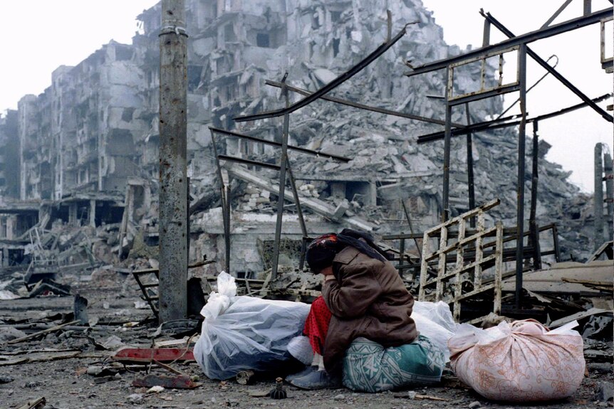A women sits slumped over and head down, behind her there is rubble everywhere and a destroyed building can be seen.
