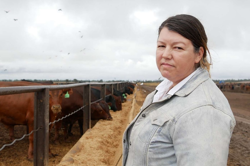 A woman stands in front of a cattle feedlot and looks at the camera, cattle eat in the background.